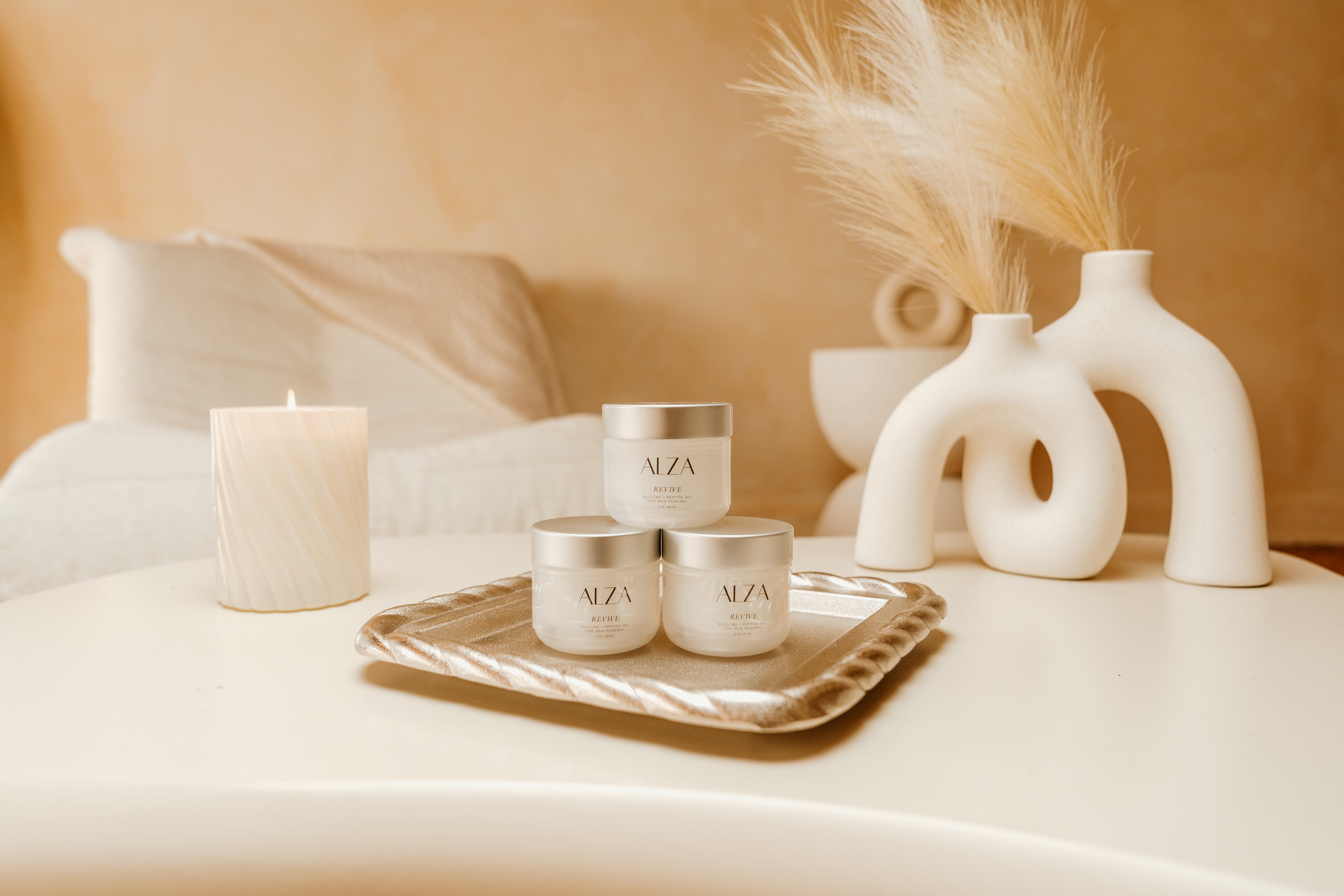 Alza skincare products on a tray with decorative vases and a candle on a neutral background